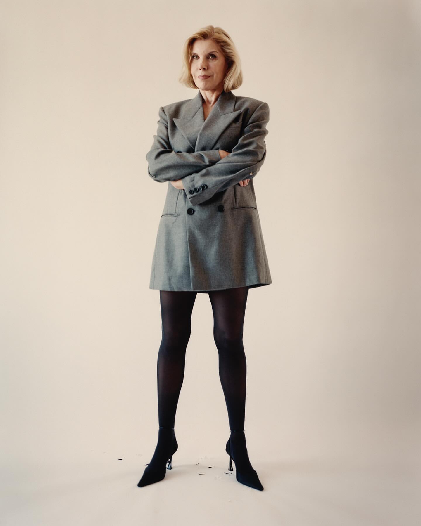 Elegant woman in gray blazer dress and black stockings posing in studio. Fashion-forward style, confident pose, neutral backdrop.