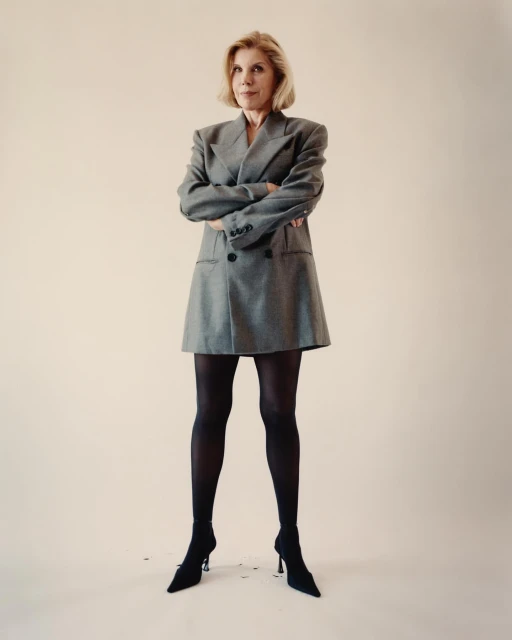 Elegant woman in gray blazer dress and black stockings posing in studio. Fashion-forward style, confident pose, neutral backdrop.