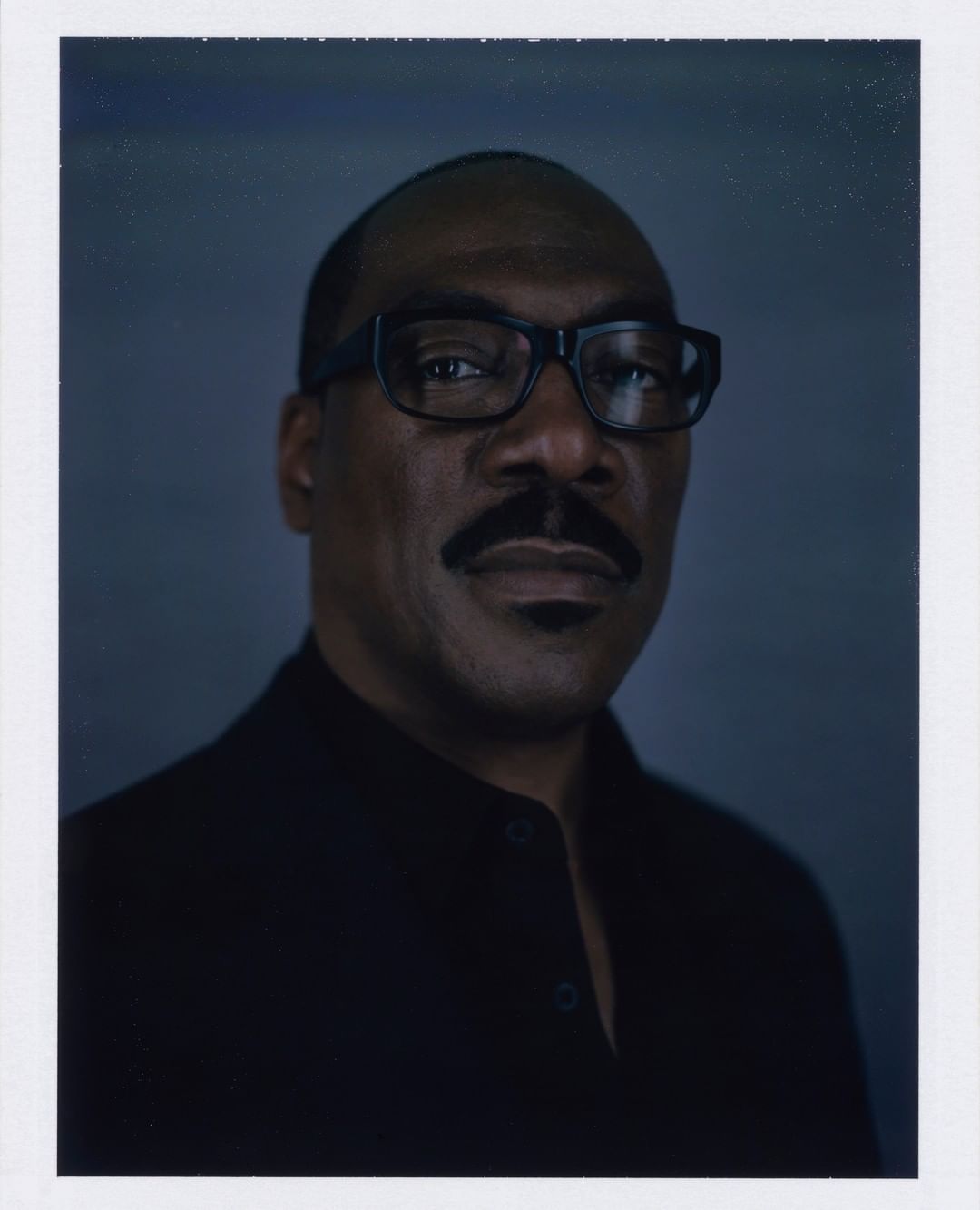 Portrait of an adult man wearing glasses, with a mustache, in a black shirt against a dark gray background with soft, diffused lighting.