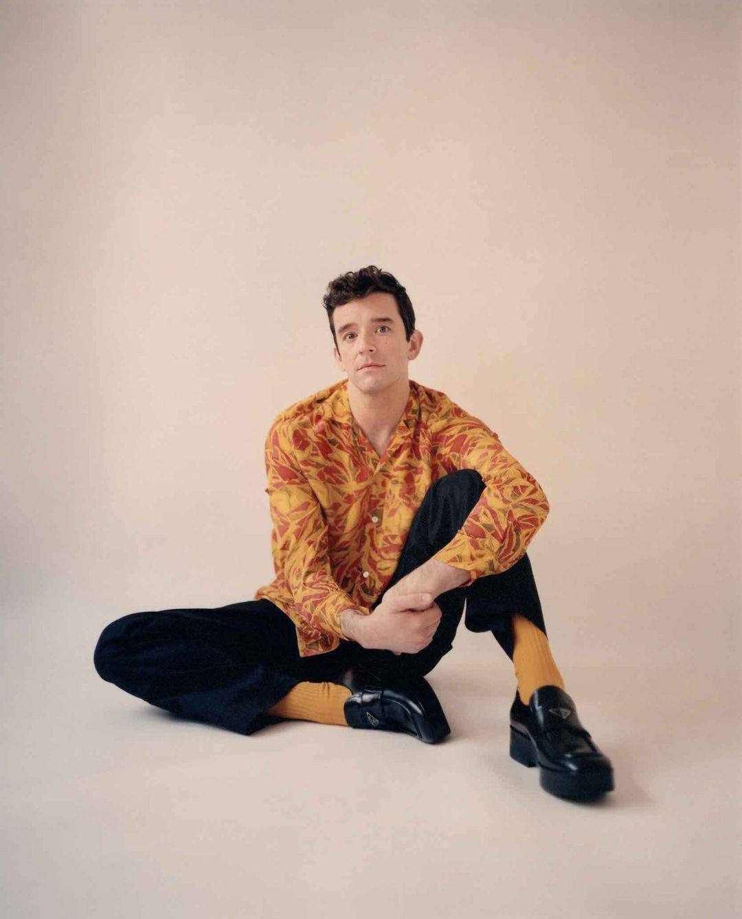 Man with dark hair seated on the floor wearing a patterned yellow shirt, dark trousers, yellow socks, and black platform loafers
