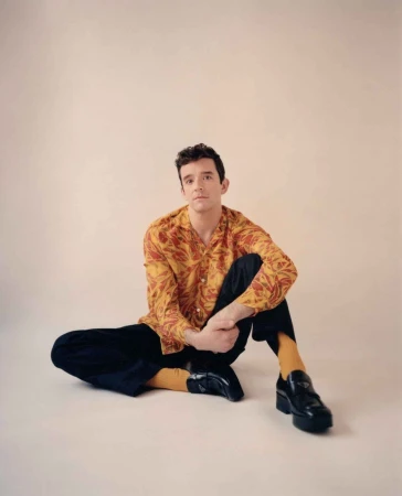 Man with dark hair seated on the floor wearing a patterned yellow shirt, dark trousers, yellow socks, and black platform loafers