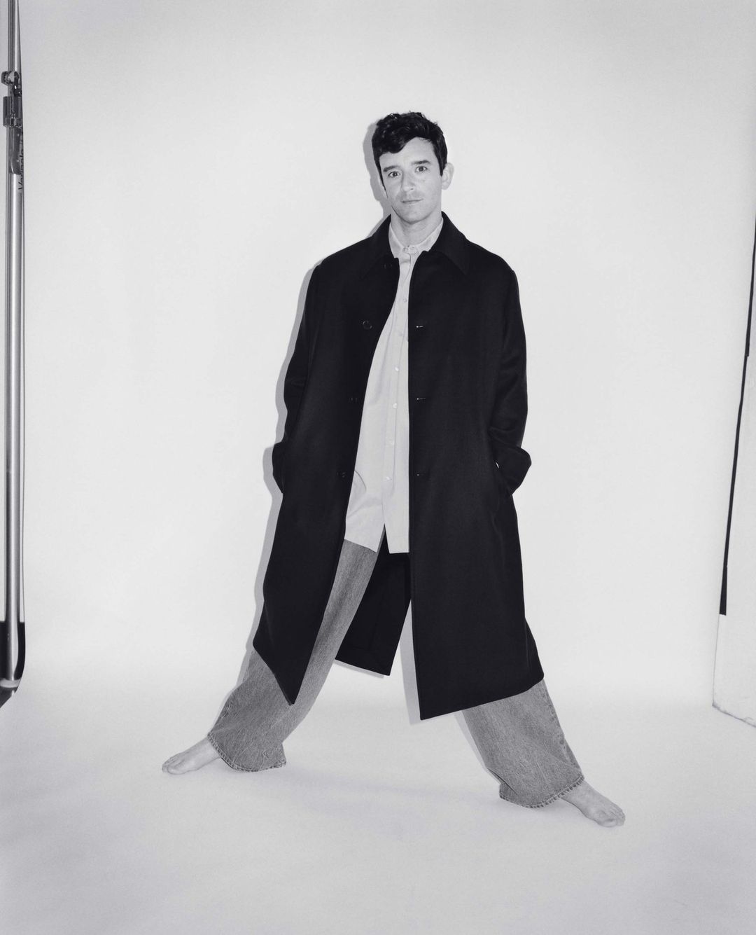 Man wearing a long black coat over a light shirt and trousers against a white seamless backdrop.