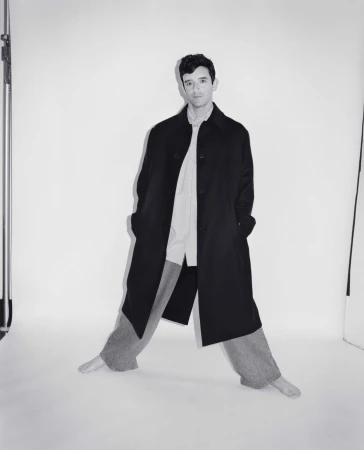 Man wearing a long black coat over a light shirt and trousers against a white seamless backdrop.