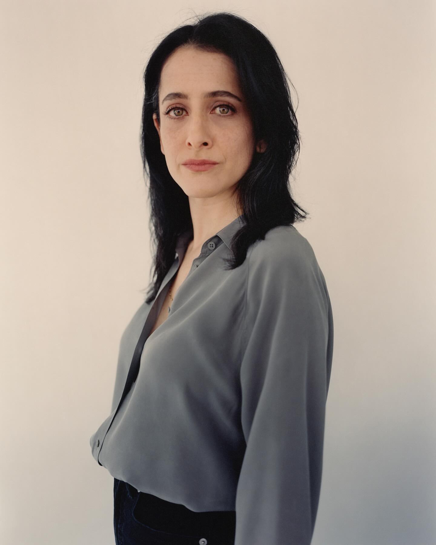 Portrait of a woman with dark hair and a grey button-down shirt, captured in a medium shot against a neutral background.