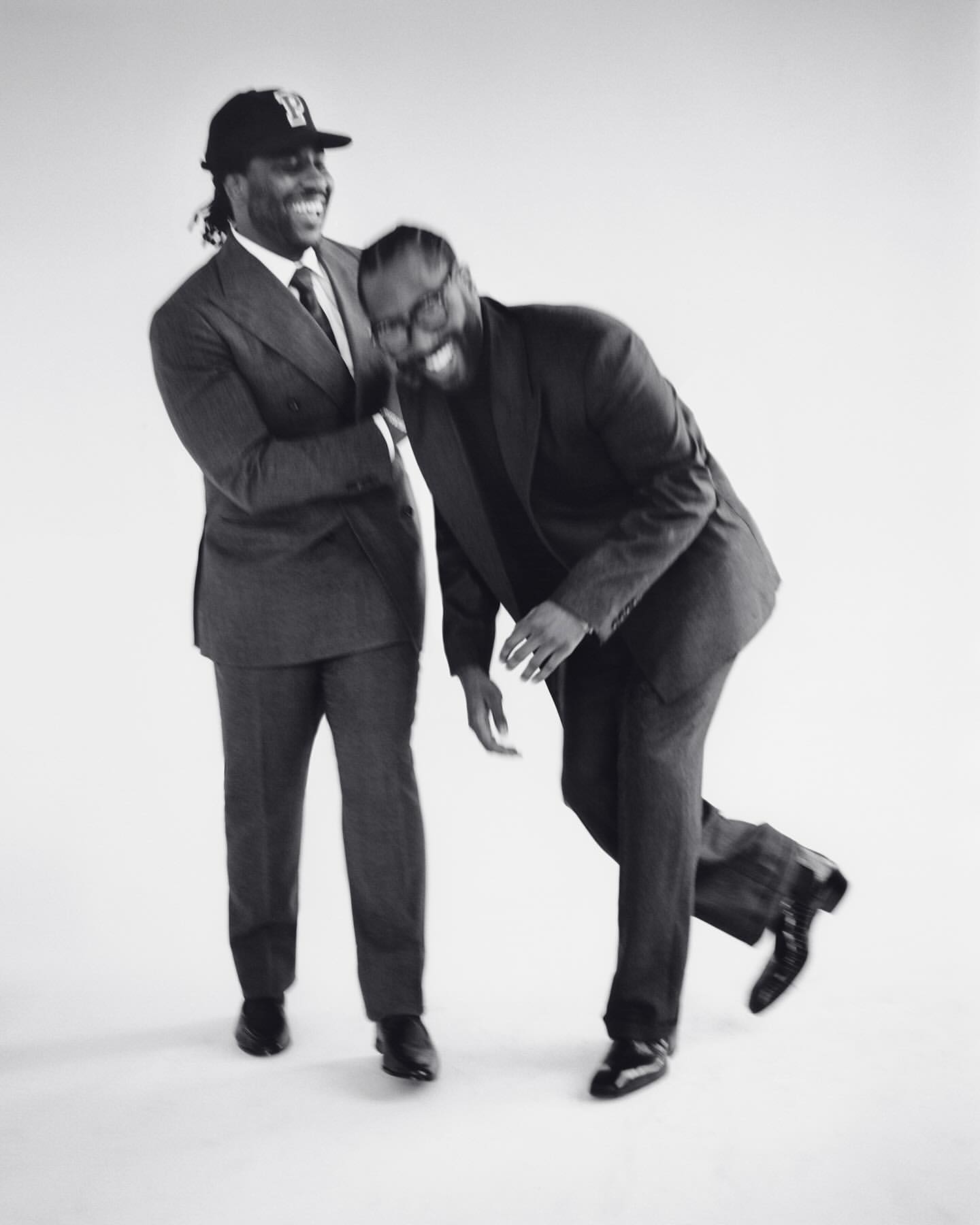 Two African-American men in suits laugh together in a studio. One wears a baseball cap, standing next to another who is leaning over.