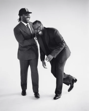 Two African-American men in suits laugh together in a studio. One wears a baseball cap, standing next to another who is leaning over.