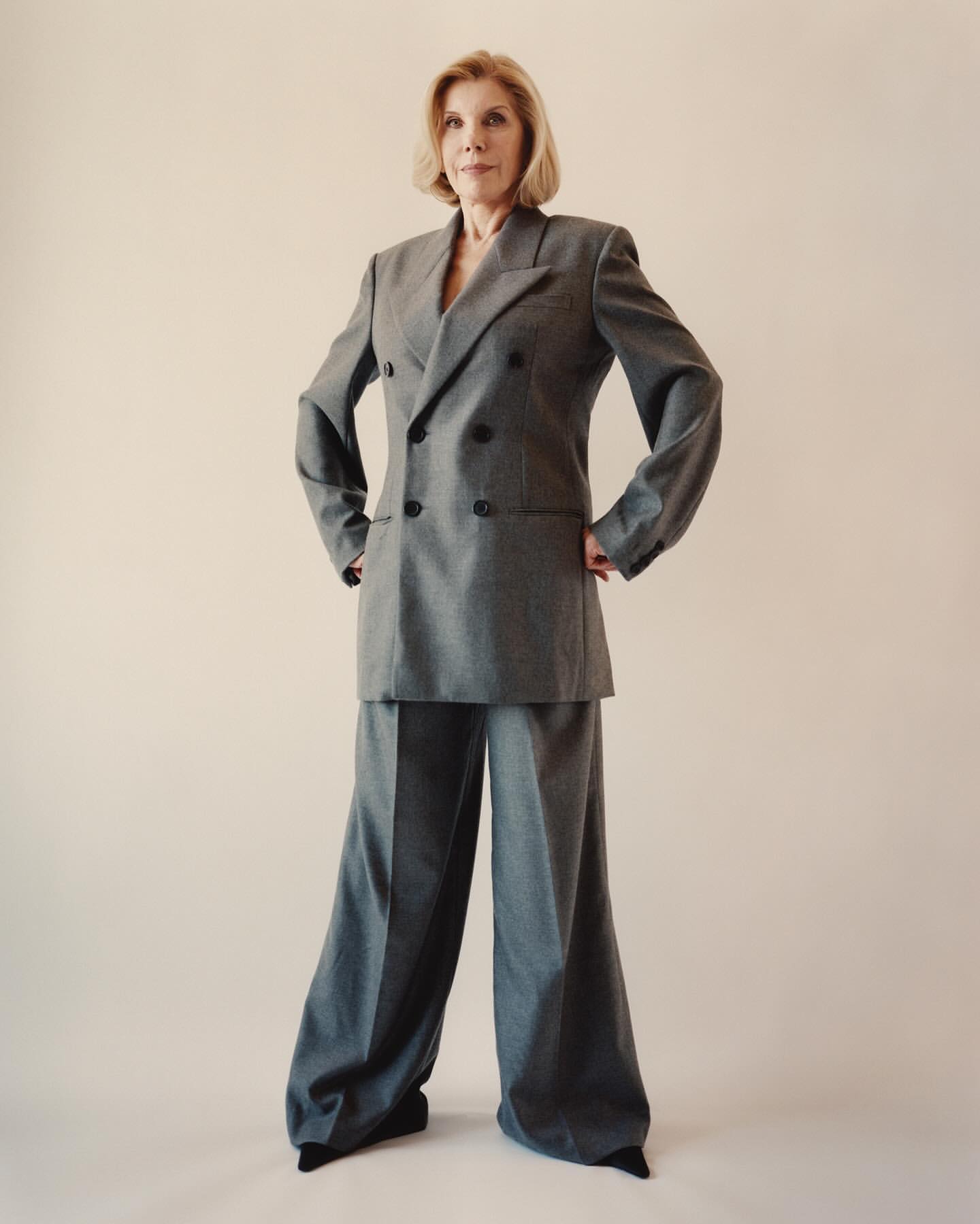 Christine Baranski poses in a grey, double-breasted suit with wide-leg pants against a neutral background, looking directly at the camera.