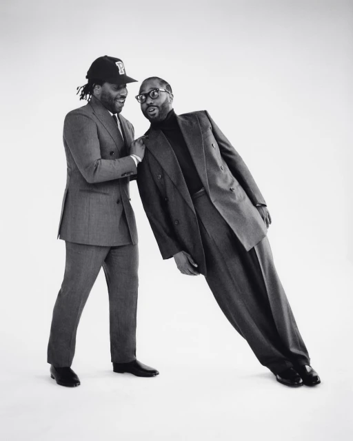 Two African American men in grey suits pose against a stark white background, with one leaning onto the other for support.