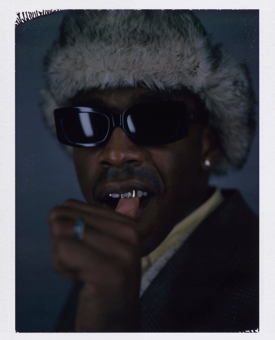Portrait of a man in sunglasses and fur hat, wearing a suit and metal grills, with hand near mouth, against a dark background.