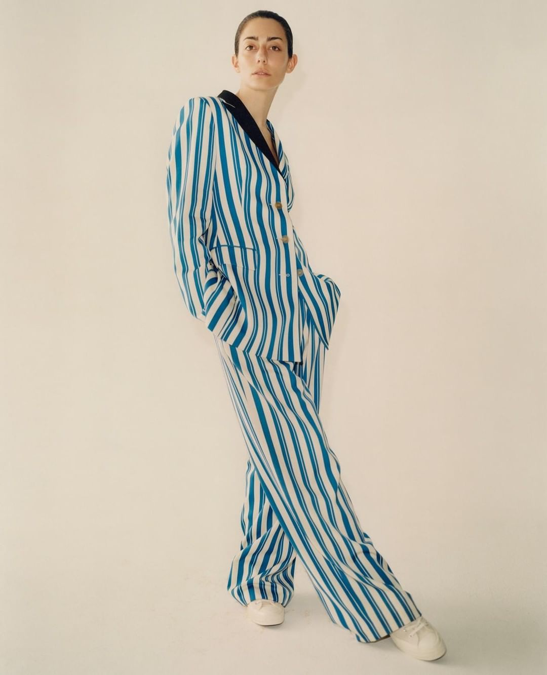 Young woman in blue-and-white striped pantsuit poses in studio setting. Vertical shot with clean, minimal aesthetic.