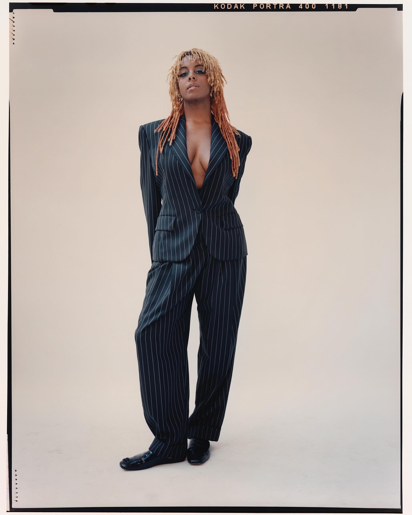 Adult female fashion model in pinstripe suit posing in studio with arms behind her back, looking at the camera. Cool studio lighting.