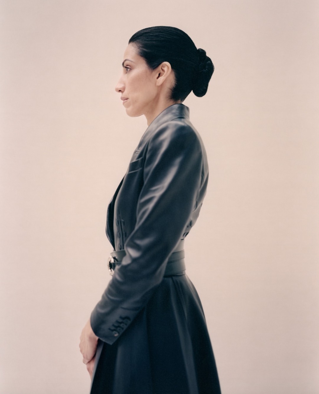 Elegant woman in profile wearing a fitted black leather dress with a matching belt, styled hair, and a light beige background.