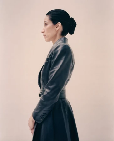 Elegant woman in profile wearing a fitted black leather dress with a matching belt, styled hair, and a light beige background.