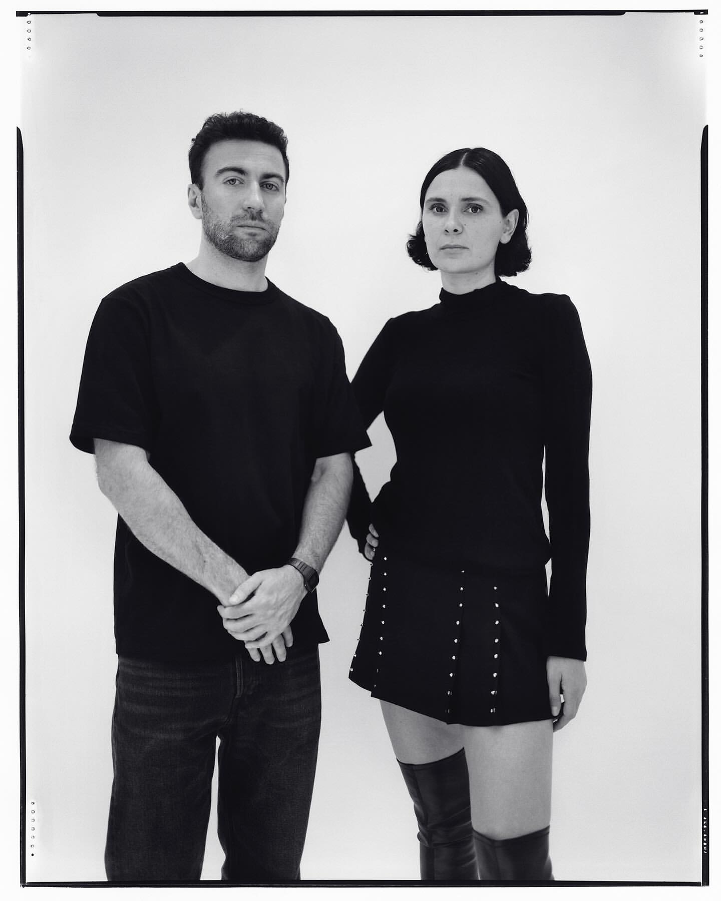 Monochrome portrait of a man in a black t-shirt and a woman in a black top with a studded miniskirt and tall boots against white seamless.