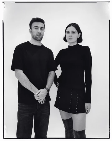 Monochrome portrait of a man in a black t-shirt and a woman in a black top with a studded miniskirt and tall boots against white seamless.