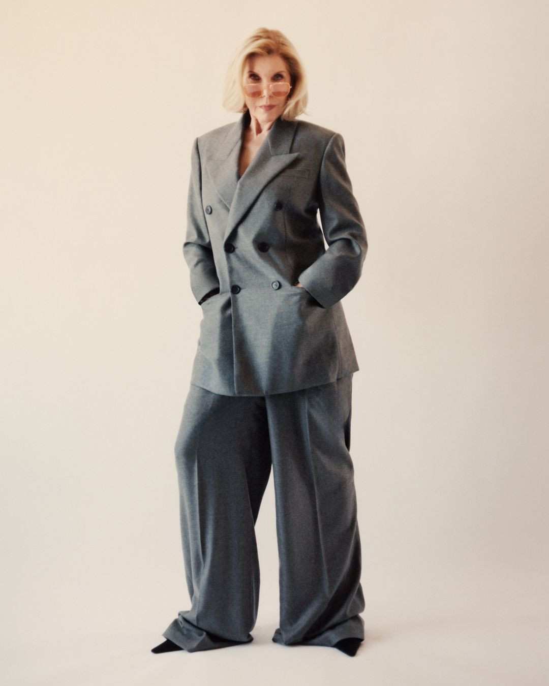 Elegant mature woman in a gray double-breasted suit with wide-leg pants and tinted glasses, confidently poses in a studio setting.