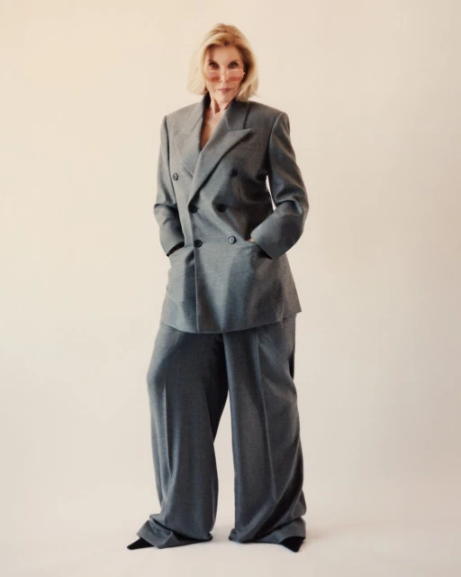 Elegant mature woman in a gray double-breasted suit with wide-leg pants and tinted glasses, confidently poses in a studio setting.