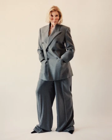 Elegant mature woman in a gray double-breasted suit with wide-leg pants and tinted glasses, confidently poses in a studio setting.