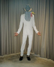 Full-length portrait of a person in a white suit and cowboy hat with silver fringe covering the face, standing before pleated curtains.