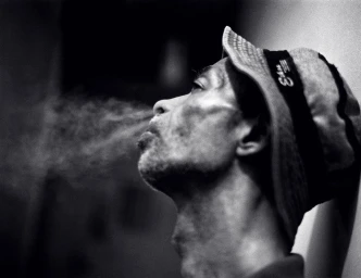 Monochrome profile portrait of a man in a bucket hat exhaling smoke against a plain background, shot by Andreas Laszlo Konrath