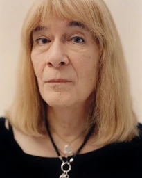 Close-up portrait of an older woman with shoulder-length blonde hair wearing a dark top and pendant necklaces