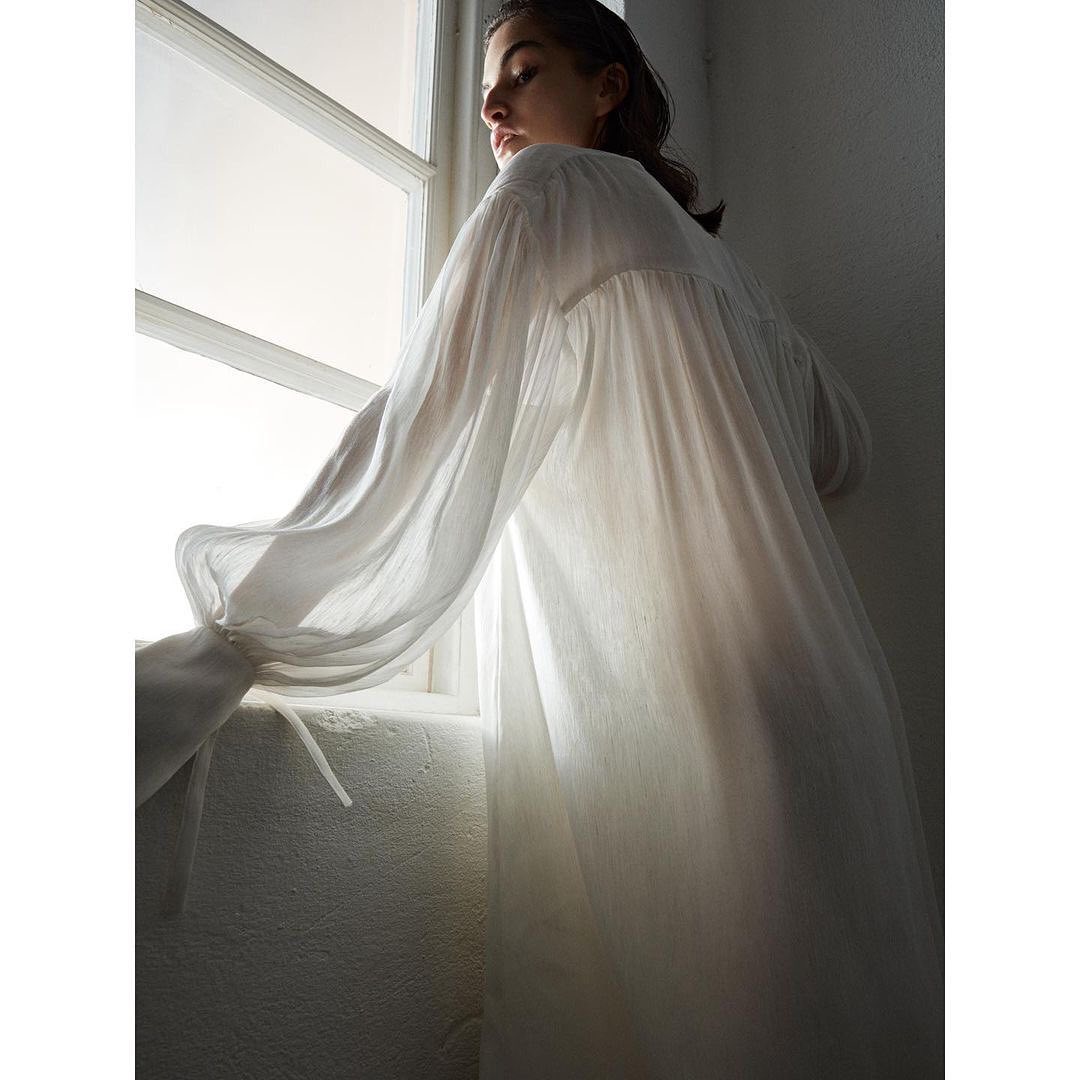 Woman in long white dress stands at window with natural light, creating a soft and serene atmosphere inside a minimally decorated room.
