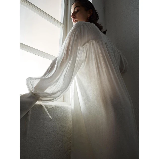 Woman in long white dress stands at window with natural light, creating a soft and serene atmosphere inside a minimally decorated room.