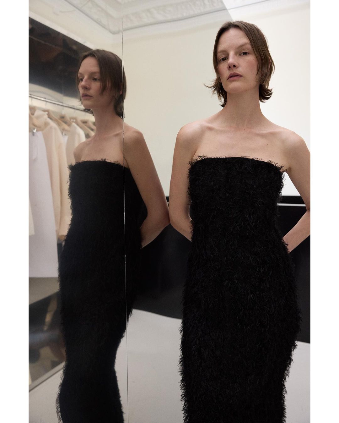 Woman in black textured strapless dress poses near a mirror inside a room with neutral tones and clothing on a rack in the background.