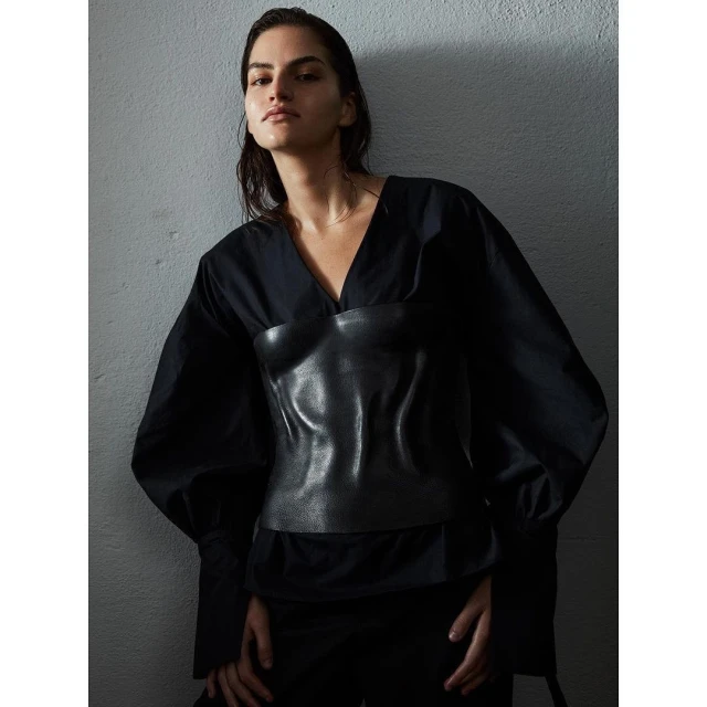 A woman poses in a black V-neck blouse with leather corset. Dark, neutral studio backdrop, dramatic lighting, sleek wet hair.