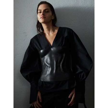 A woman poses in a black V-neck blouse with leather corset. Dark, neutral studio backdrop, dramatic lighting, sleek wet hair.