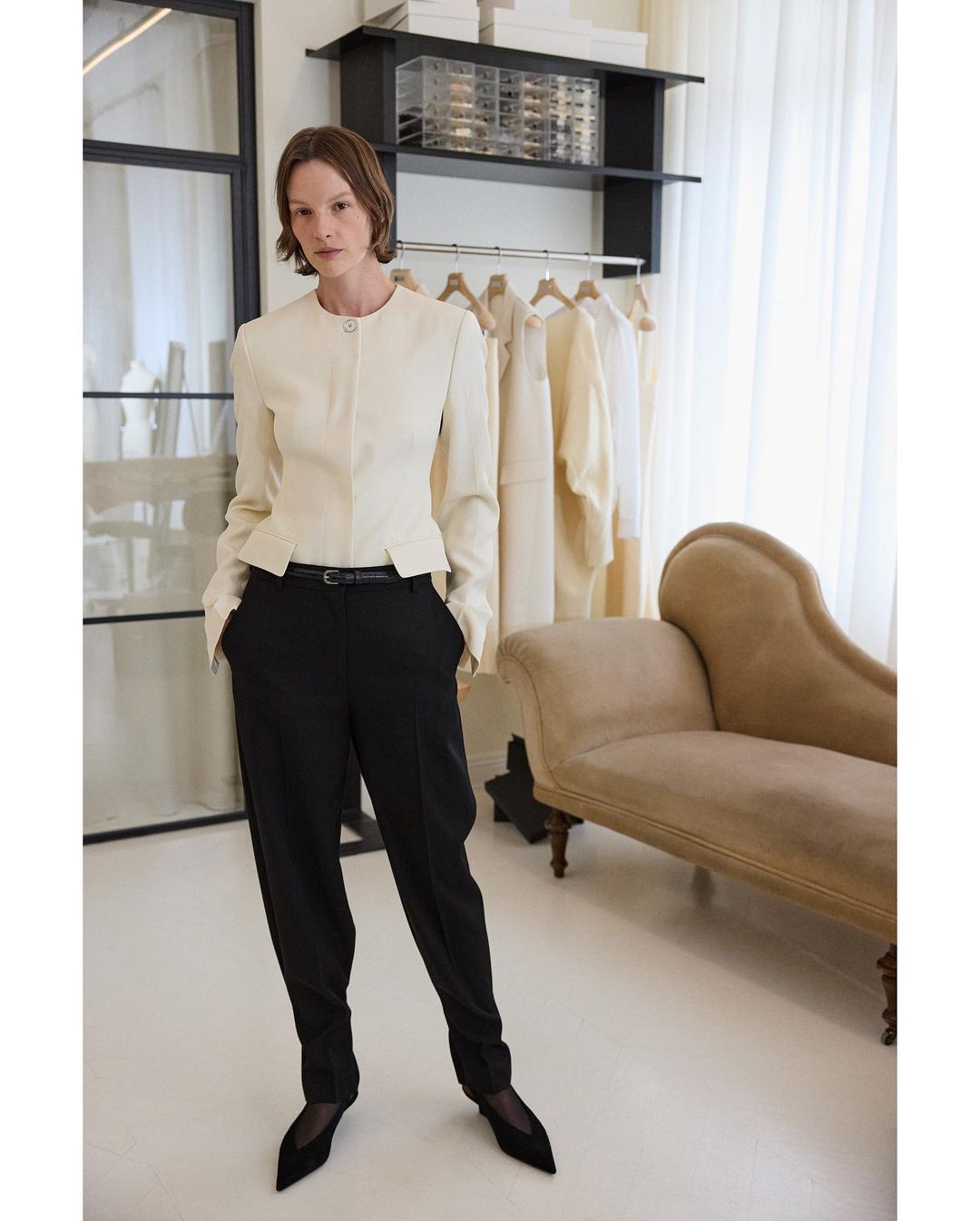 Elegant woman posing in a cream jacket and black pants. Fashion shot in a light interior with a chaise lounge and clothing rack.