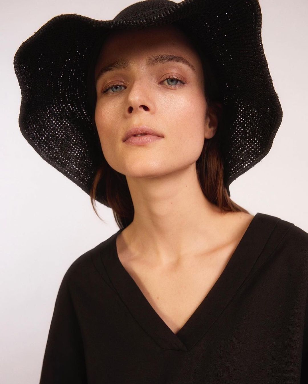 Close-up portrait of a woman wearing a black V-neck top and a black straw sun hat against a plain off-white studio backdrop.