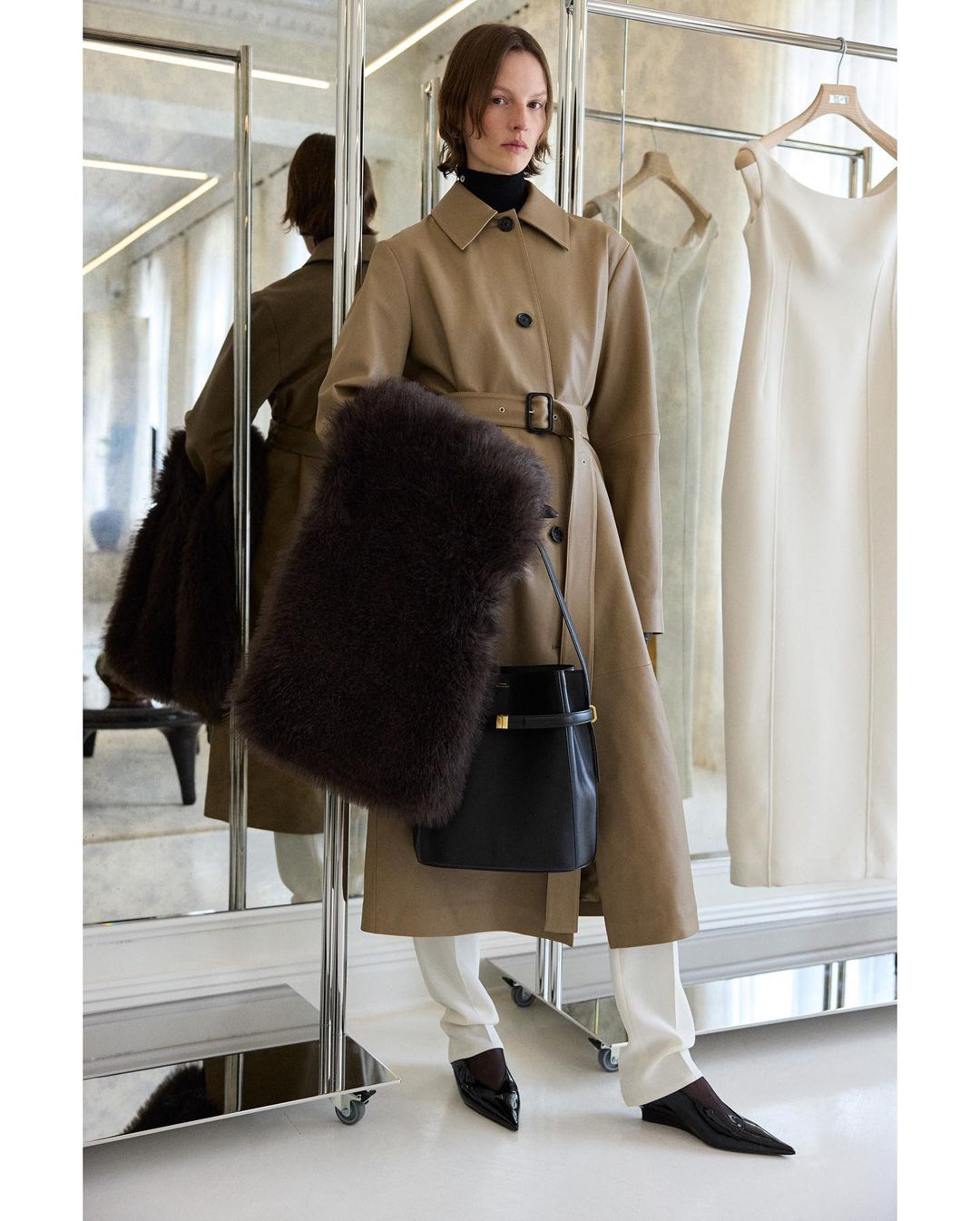 Fashion shot featuring an adult female wearing a tan trench coat, white pants, and a black shoulder bag in an indoor setting.