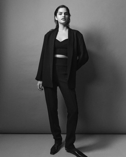 Black and white full shot of a young woman in a black blazer, top, and trousers posing against a gray background in a photo studio.