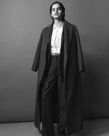 Monochrome fashion portrait of a model wearing a long wool coat, button-up shirt, and trousers against a grey backdrop