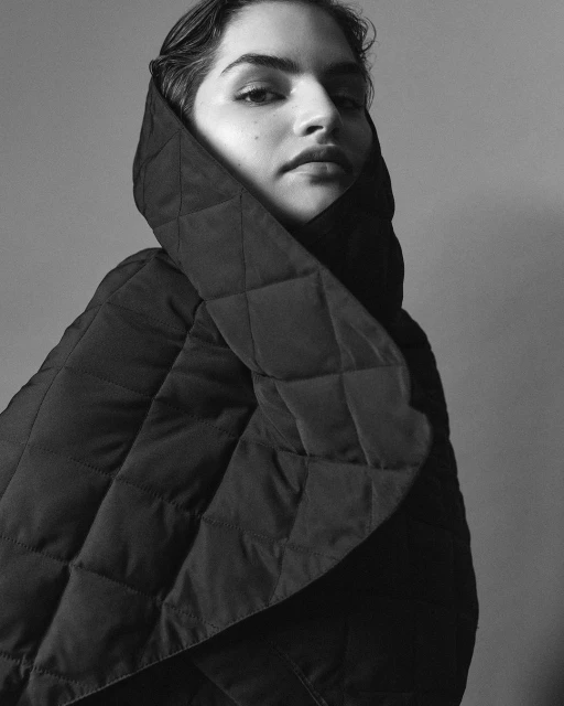 Black and white portrait of a young adult female with medium skin tone wearing a dark, quilted jacket with a high, puffy collar.