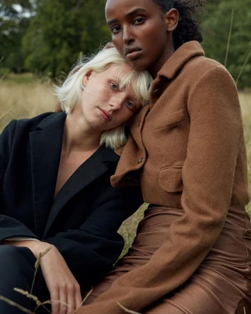 Two women posing outdoors in a grassy field, one resting her head gently on the shoulder of the other.