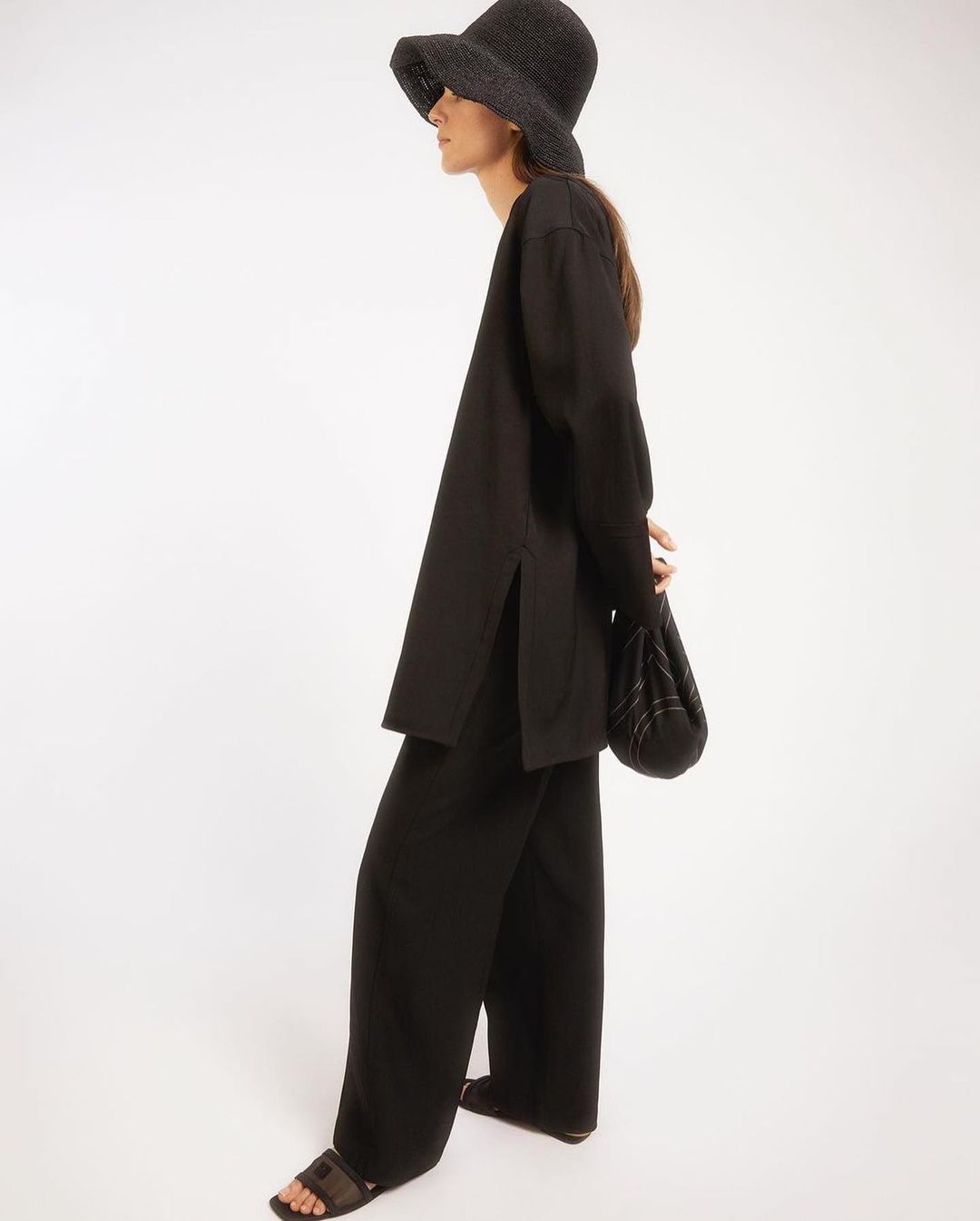Fashionable woman in black wide-leg pants, tunic top, woven hat, and bag on a white background in a studio shot.