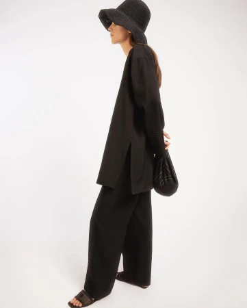 Fashionable woman in black wide-leg pants, tunic top, woven hat, and bag on a white background in a studio shot.