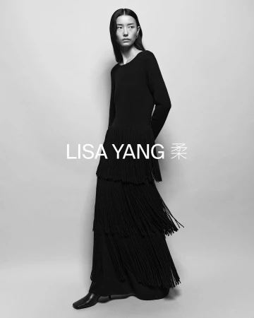 Black and white fashion shot of an Asian woman in a long-sleeved black dress with layered fringe skirt, wearing square-toed boots.