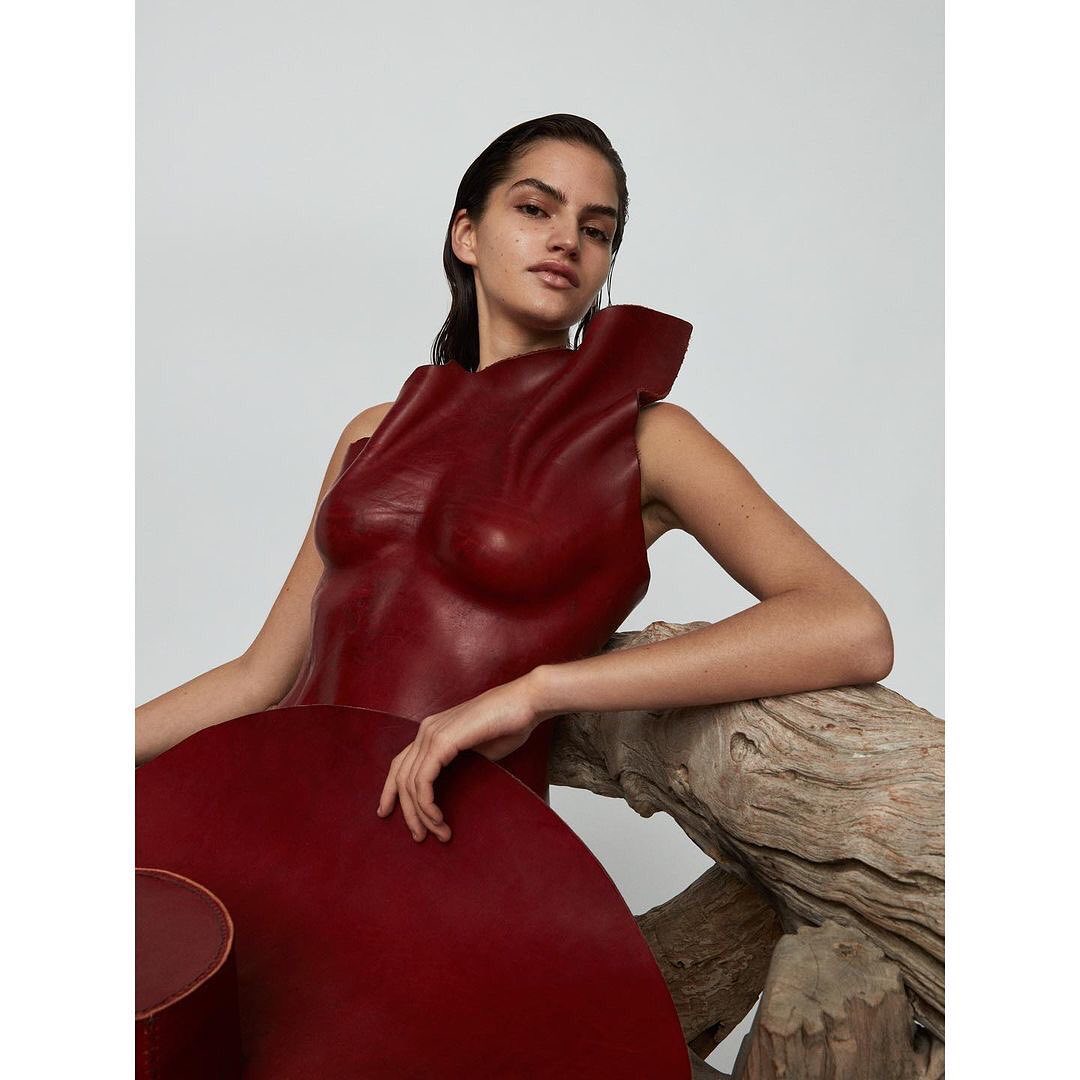 Brunette woman in a sculptural red dress poses with weathered wood against a plain background in a studio-lit fashion shot.