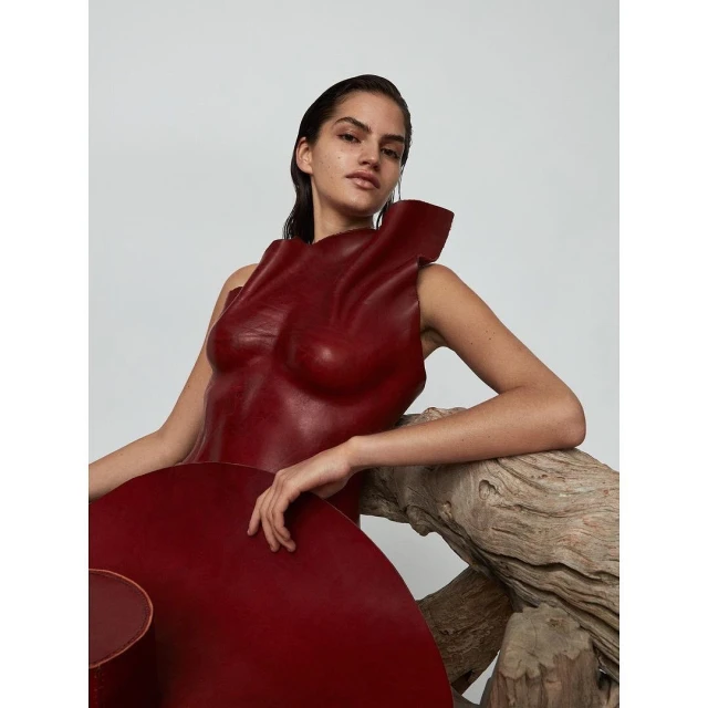 Brunette woman in a sculptural red dress poses with weathered wood against a plain background in a studio-lit fashion shot.