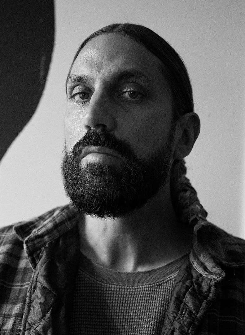 Monochrome portrait of an adult male with a beard and long hair, wearing a flannel shirt, gazing directly at the camera in a studio setting.
