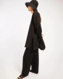 Model in black tunic and trousers wearing a wide-brim hat holding a small dark bag against a white seamless backdrop.