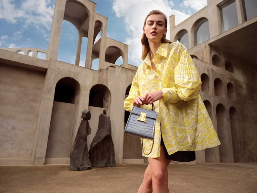 A fair-skinned woman in a yellow patterned blouse poses with a grey handbag against an architectural backdrop with an overcast sky.