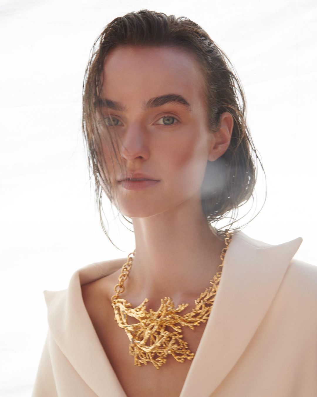 Close-up studio shot of a young adult female model with golden statement necklace, wearing off-white jacket, and hair partially wet.