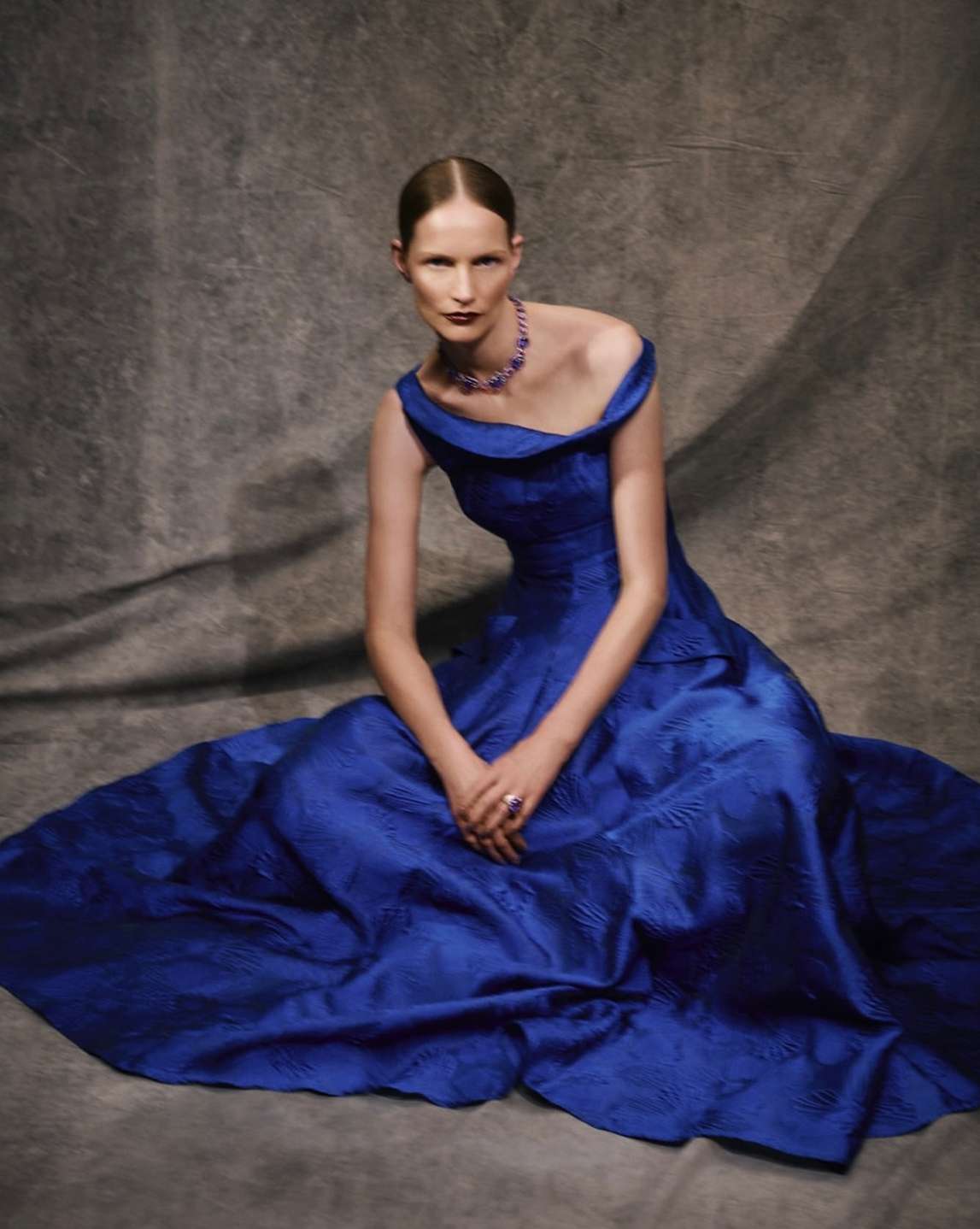 Elegant adult woman in blue off-the-shoulder gown and jewel necklace sitting against a grey textured backdrop in a fashion portrait.