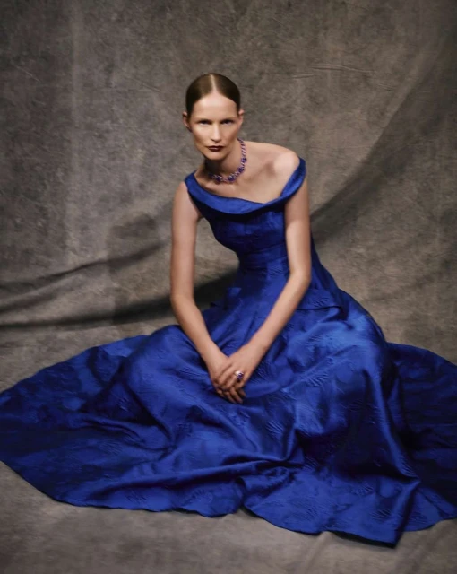 Elegant adult woman in blue off-the-shoulder gown and jewel necklace sitting against a grey textured backdrop in a fashion portrait.
