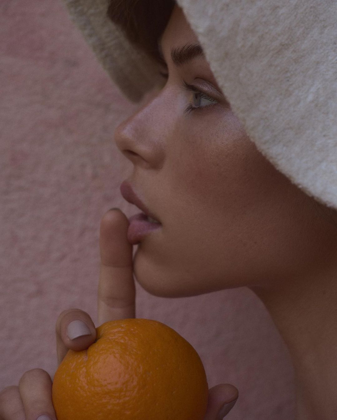 Close-up portrait of a woman in a hat holding an orange, with her finger to her lips against a textured light pink background.