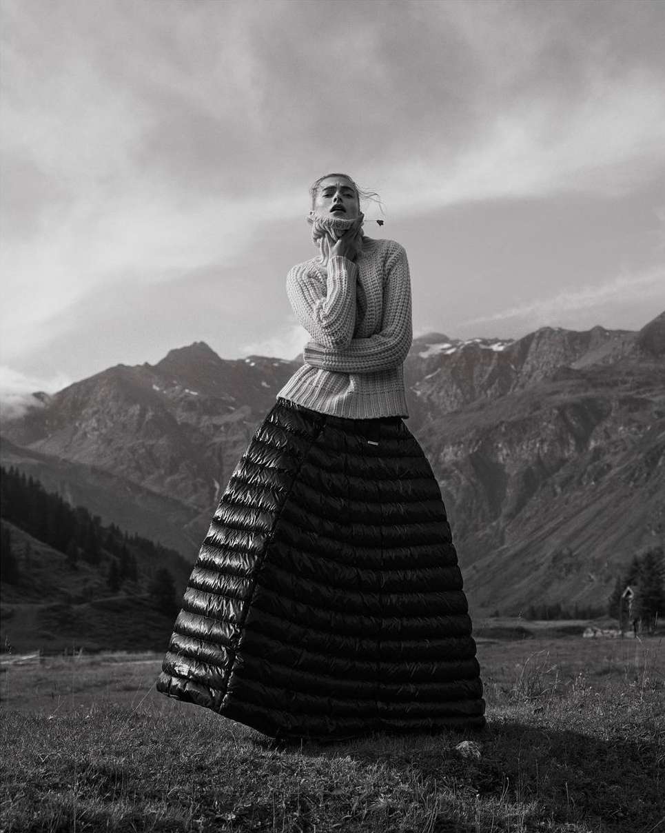 Monochrome fashion shot of a woman in a sweater and quilted skirt, posing in a field with mountains and a cloudy sky in the background.